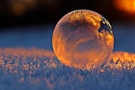 Close-up shot of a frozen bubble with warm reflections resting on a snowy surface at twilight.